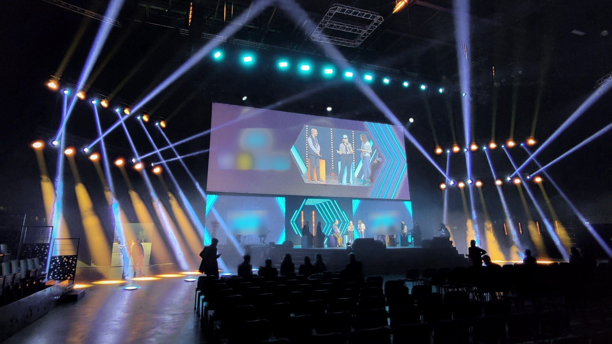 Une salle en cours de montage à contre-jour : un technicien règle un projecteur, deux personnes alignent les chaises.