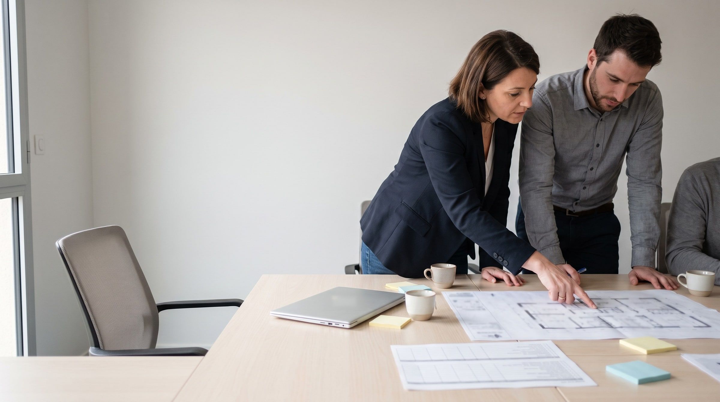 Deux personnes penchées sur des plans imprimés posés sur une table de réunion, en lumière du jour.